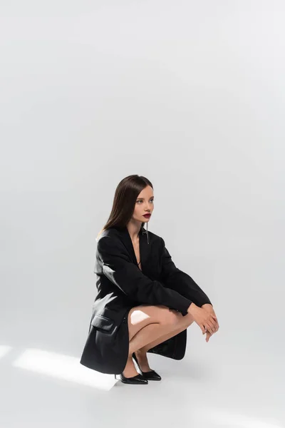 Сексуальная женщина в черном пиджаке и туфлях на высоких каблуках смотрит в сторону, позируя на сером — стоковое фото
