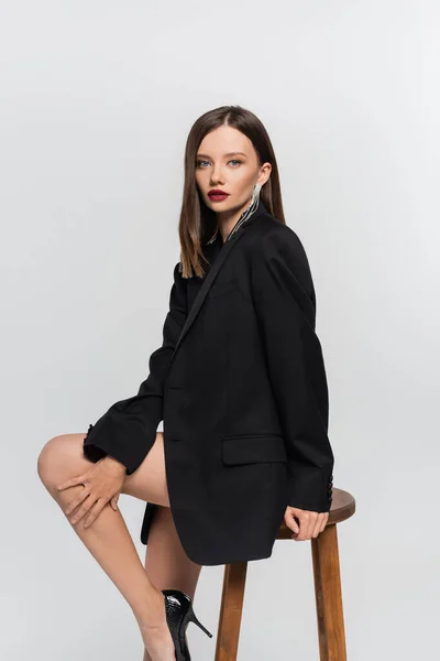 Seductive brunette woman with naked legs, wearing black blazer, looking at camera on high stool isolated on grey — Stock Photo