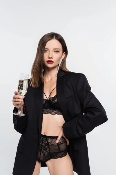 Femme brune avec verre de champagne posant en sous-vêtements en dentelle et blazer noir isolé sur gris — Photo de stock