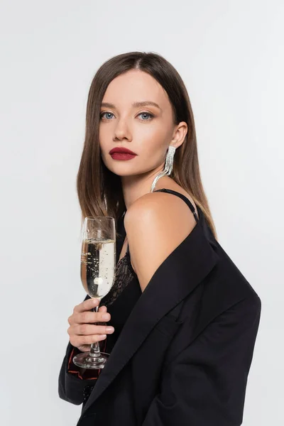 Sexy morena mulher no preto blazer olhando para câmera enquanto segurando copo de champanhe isolado no cinza — Fotografia de Stock
