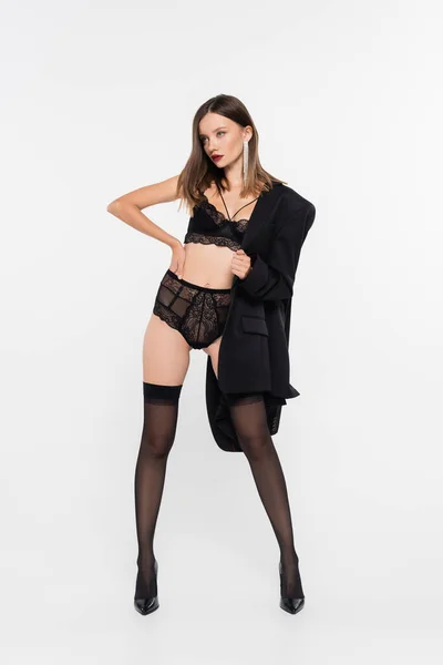 Mulher sedutora em lingerie de renda e meias pretas posando com a mão no quadril e blazer preto no cinza — Fotografia de Stock