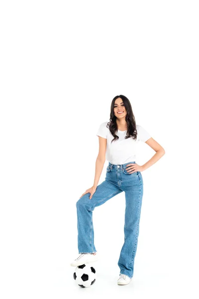 Full length view of happy woman in jeans stepping on soccer ball while standing with hand on hip on white — стоковое фото