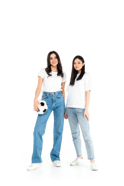 Full length view of brunette woman in jeans holding ball near asian friend on white - foto de stock