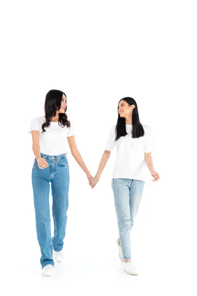 Visão de comprimento total de mulheres inter-raciais felizes em jeans de mãos dadas enquanto caminhava sobre branco — Fotografia de Stock