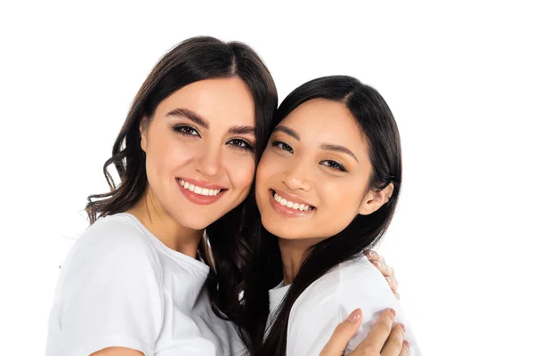 Happy brunette woman embracing and looking at camera with asian friend isolated on white — Stock Photo