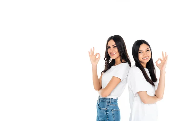 Brunette multiethnic women showing okay signs and smiling at camera isolated on white — стоковое фото