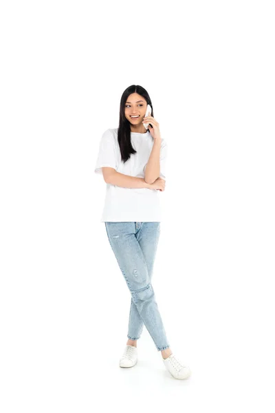 Full length view of happy asian woman in t-shirt and jeans talking on smartphone on white — стоковое фото