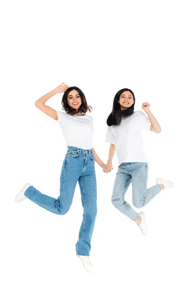 Overjoyed interracial women in jeans holding hands while levitating isolated on white - foto de stock