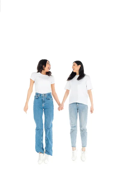Full length view of astonished interracial women holding hands and looking at each other while levitating isolated on white - foto de stock
