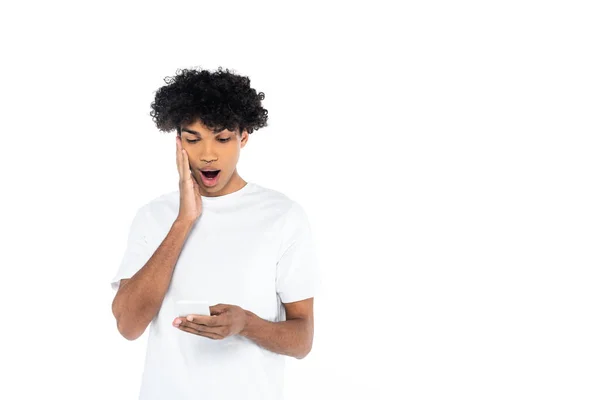 Шокований афроамериканський чоловік торкається обличчя, дивлячись на мобільний телефон ізольовано на білому — стокове фото