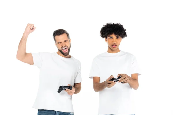 KYIV, UKRAINE - DECEMBER 5, 2021: excited man showing win gesture while gaming with upset african american friend isolated on white — Stock Photo