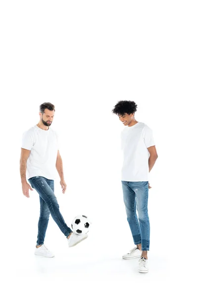 Uomo afroamericano sorridente in piedi con le mani dietro la schiena vicino amico che gioca con il pallone da calcio su bianco — Foto stock