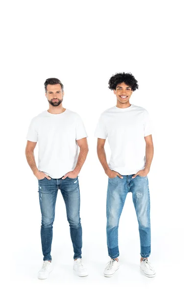 Full length view of interracial men smiling at camera while standing with hands in pockets on white - foto de stock