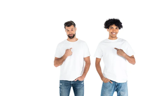 Fröhliche multiethnische Männer, die mit den Händen in den Taschen stehen und auf sich selbst zeigen, isoliert auf weiß — Stockfoto