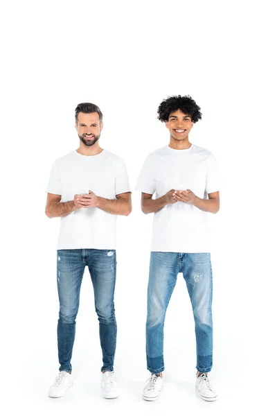 Vista a figura intera di uomini felici multietnici in t-shirt e jeans che chattano su smartphone su bianco — Foto stock