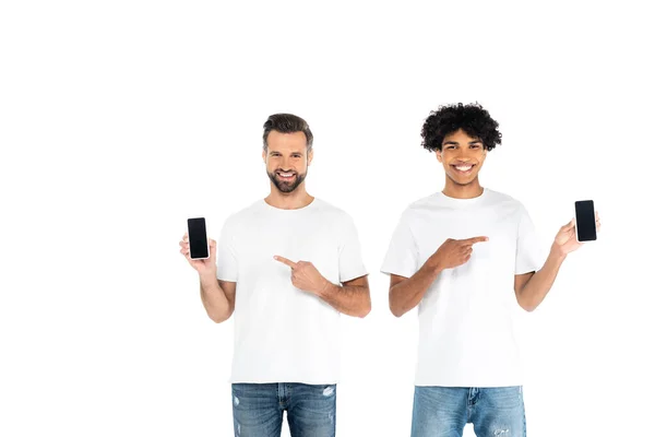 Sonrientes hombres multiétnicos señalando con los dedos a los teléfonos inteligentes con pantalla en blanco aislado en blanco - foto de stock