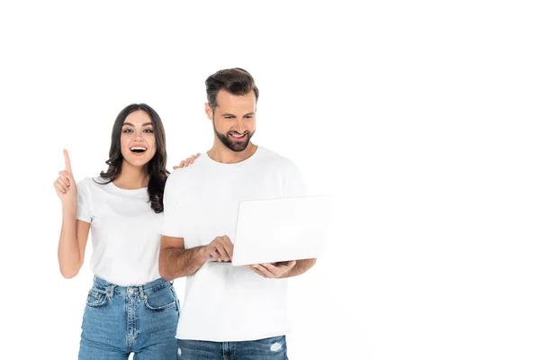 Mujer excitada señalando con el dedo cerca de hombre sonriente con el ordenador portátil aislado en blanco - foto de stock