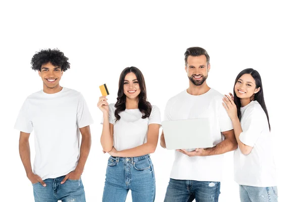 Sonriente mujer sosteniendo tarjeta de crédito cerca de hombre con portátil y amigos interracial aislados en blanco - foto de stock
