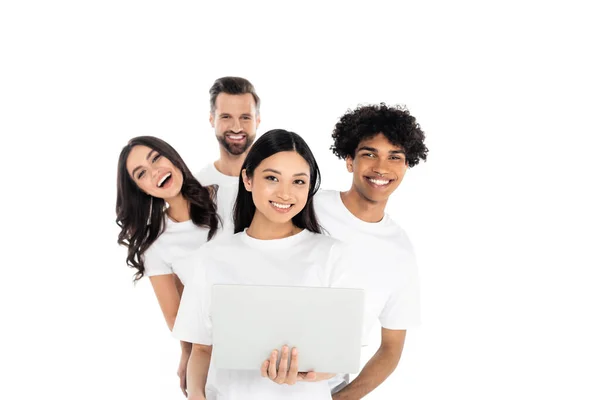 Alegres amigos multiétnicos mirando a la cámara cerca de mujer asiática con portátil aislado en blanco - foto de stock