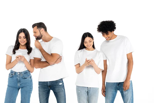 Веселі міжрасові жінки обмінюються повідомленнями на мобільних телефонах поблизу цікавих чоловіків ізольовані на білому — стокове фото