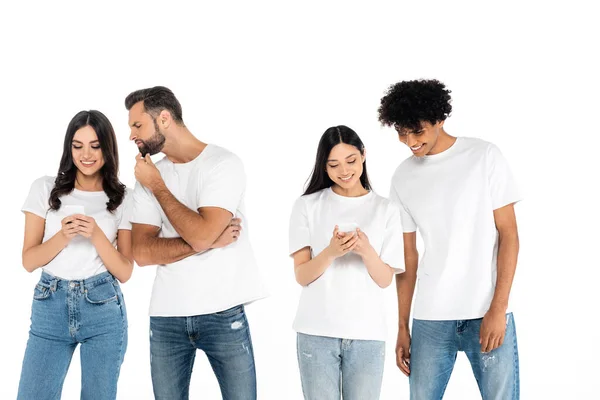 Alegre interracial las mujeres mensajería en los teléfonos celulares cerca curiosos hombres aislados en blanco - foto de stock