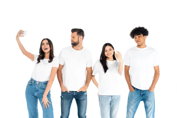 Hombres escépticos de pie con las manos en los bolsillos cerca de las mujeres interracial felices tomando selfie aislado en blanco - foto de stock