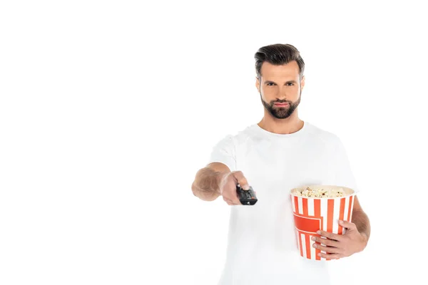 Bearded man clicking tv channels on remote controller while holding big bucket of popcorn isolated on white — Stockfoto