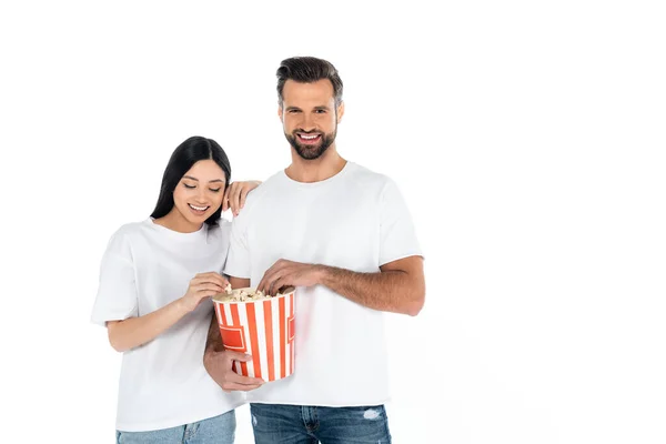 Smiling man holding popcorn bucket near happy asian woman isolated on white — стоковое фото