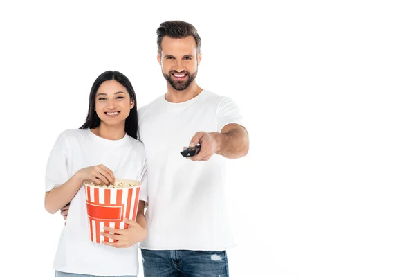 Zufriedener Mann klickt Fernsehkanäle nahe asiatische Frau mit Eimer Popcorn isoliert auf weiß — Stockfoto