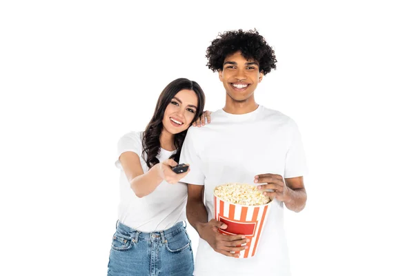 Donna sorridente cliccando canali tv vicino felice uomo africano americano con popcorn isolato su bianco — Foto stock