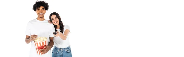 Joyful woman clicking tv channels near african american man with bucket of popcorn isolated on white, banner — Stock Photo