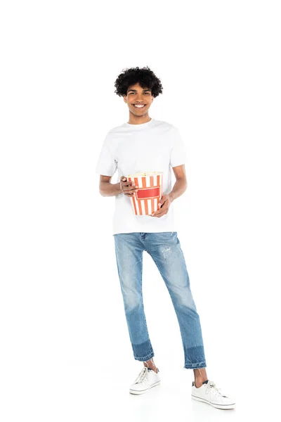 Ganzkörperansicht eines glücklichen afrikanisch-amerikanischen Mannes in Jeans, der einen Eimer Popcorn auf weiß hält — Stockfoto