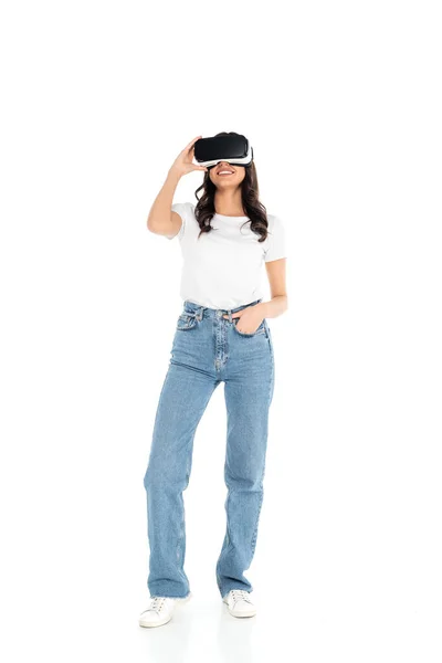 Full length view of happy woman standing with hand in pocket while gaming in vr headset on white — Photo de stock