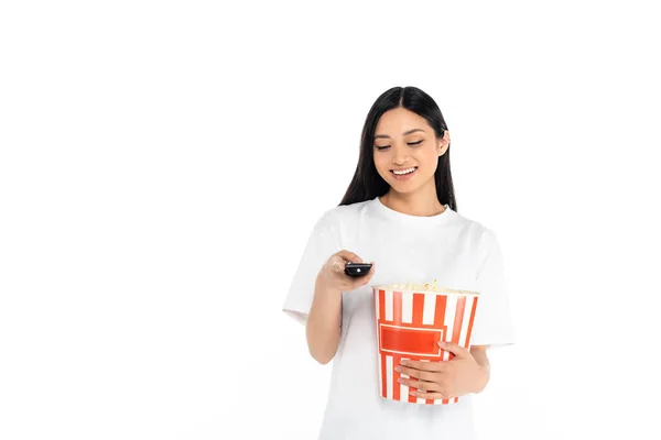 Happy asian woman clicking tv channels while holding large popcorn bucket isolated on white — Stock Photo