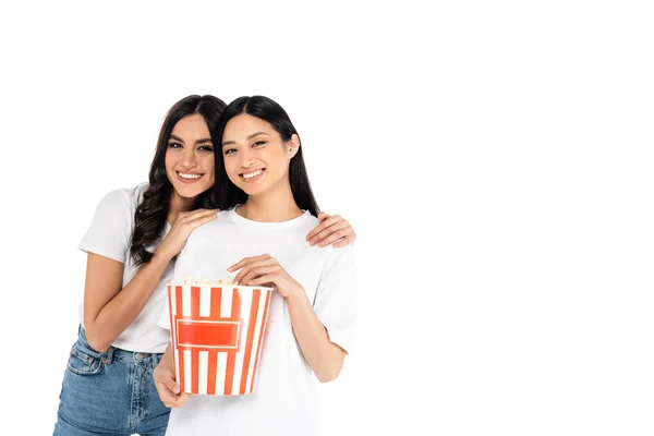 Happy woman embracing asian friend holding big bucket of popcorn isolated on white — Fotografia de Stock