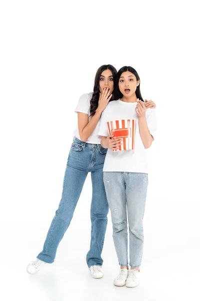 Shocked woman covering mouth with hand while watching movie with asian friend holding bucket of popcorn on white — стоковое фото