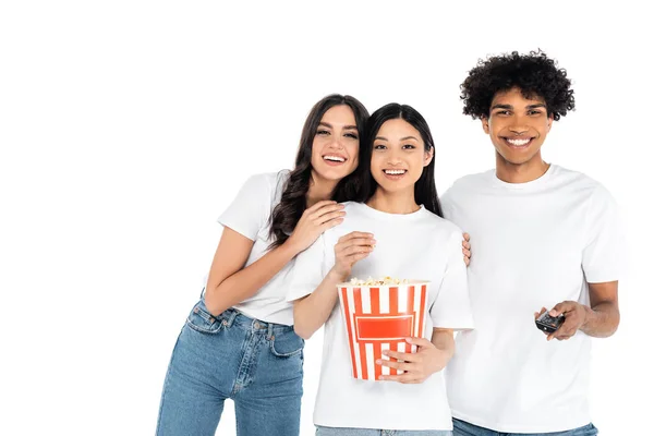 Sorridente uomo africano americano cliccando canali tv vicino felice donne interrazziali con secchio di popcorn isolato su bianco — Foto stock