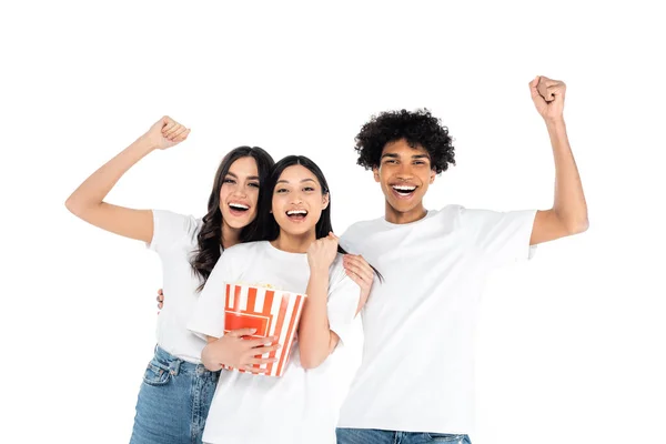 Excited multiethnic friends showing triumph gesture near bucket of popcorn isolated on white — Stockfoto