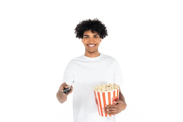 Alegre afroamericano hombre con un cubo de palomitas de maíz haciendo clic en canales de televisión aislados en blanco - foto de stock