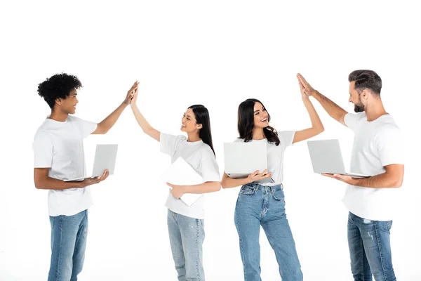 Cheerful multicultural friends with laptops giving high five isolated on white — Stock Photo