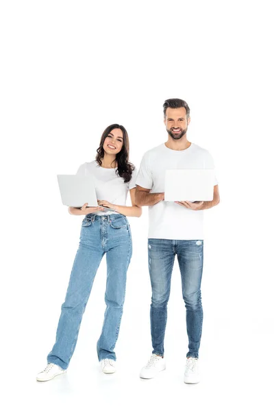 Full length view of happy couple in jeans using laptops while looking at camera on white — Fotografia de Stock