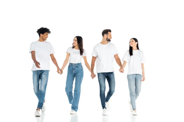 Full length view of interracial friends in jeans looking at each other and holding hands while walking on white — Stock Photo