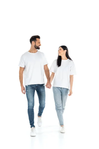 Full length view of happy interracial couple looking at each other and holding hands while walking on white — стоковое фото