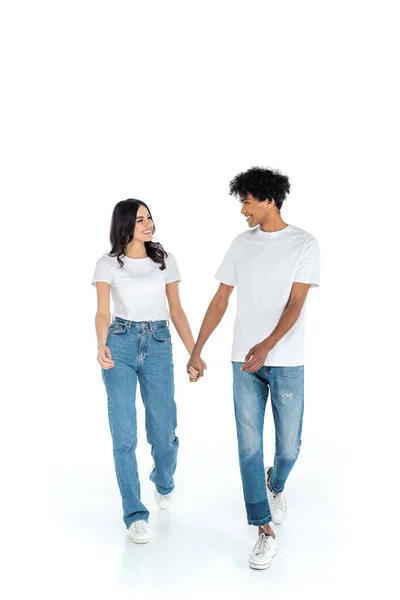 Full length view of happy interracial couple holding hands and looking at each other while walking on white — Fotografia de Stock