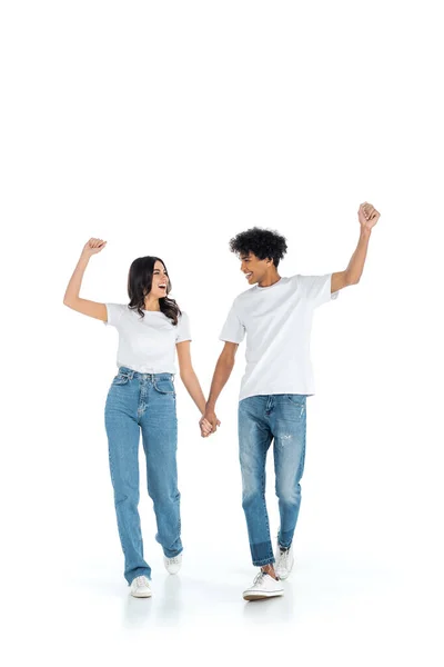 Excited multiethnic couple showing win gesture while holding hands and walking on white — Stock Photo