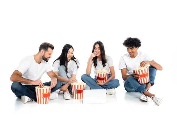 Sonrientes amigos multiétnicos comiendo palomitas de maíz mientras ven películas en el portátil en blanco - foto de stock