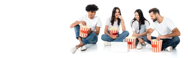 Amigos multiétnicos alegres viendo la película en el ordenador portátil y comer palomitas de maíz mientras se sienta en blanco, bandera - foto de stock