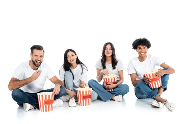 Joyful multicultural friends sitting with popcorn buckets and watching movie on white — Stock Photo