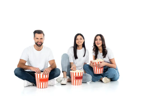 Amici multietnici felici in jeans seduti vicino a secchi di popcorn su bianco — Foto stock