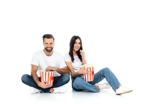 Visão de comprimento total de casal feliz em jeans comer pipocas enquanto sentado em branco — Fotografia de Stock